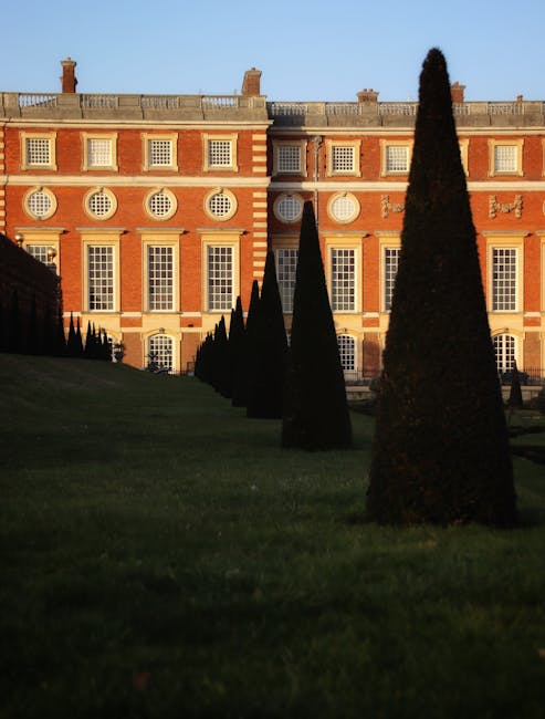 A historic brick mansion with large, evenly spaced sash windows and decorative white trim on its facade, situated in a well-maintained formal garden. In the foreground, neatly trimmed conical topiary 